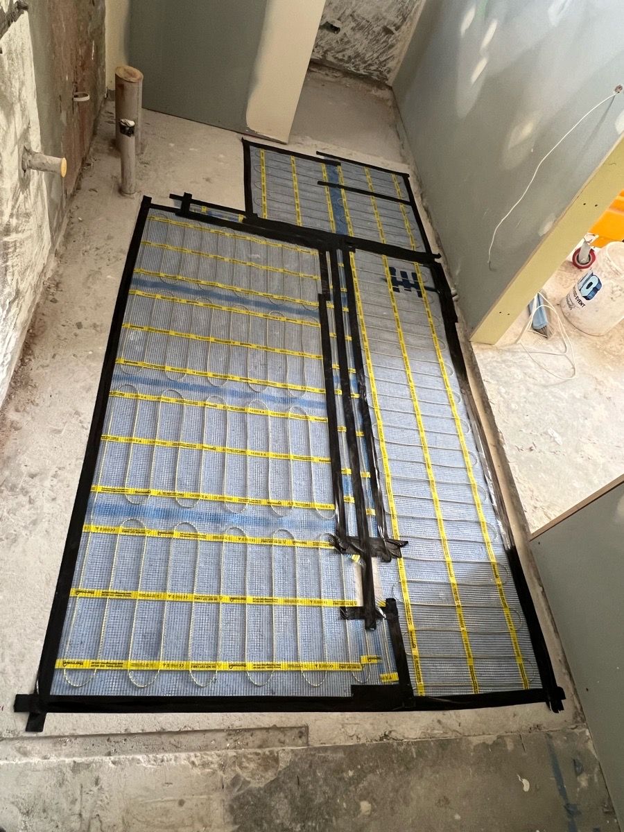 Electric heating mat laid on a bathroom floor, cut around walls and pipe penetrations, with cold leads gathered in the middle